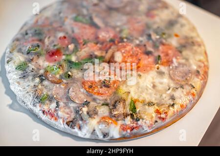 Pizza suprêmes toute entière congelée avec une croûte mince de plastique enveloppée sur la table avec des saupoudrés de fromage mozzarella et de la sauce tomate rouge, des tranches de pepperoni et Banque D'Images