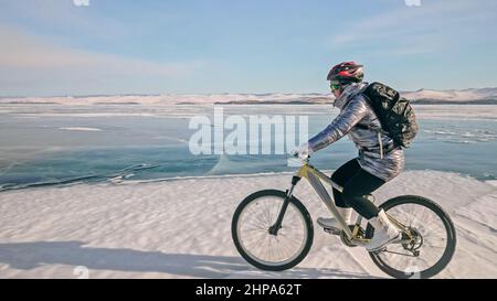 Une femme est à vélo sur la glace. La fille est vêtue d'une veste en duvet argenté, d'un sac à dos de cyclisme et d'un casque. Glace du lac Baïkal gelé. Pneus sur bik Banque D'Images