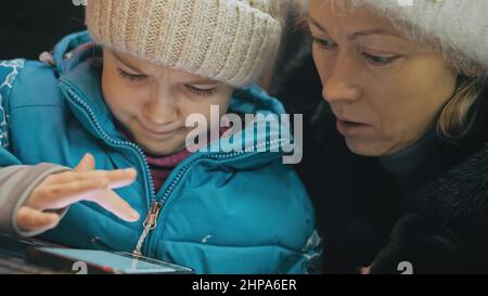 Belle mère et fille caucasienne utilisant un smartphone assis dans un café d'hiver en veste, chapeau. Jeune fille avancée montre et explique à maman Banque D'Images