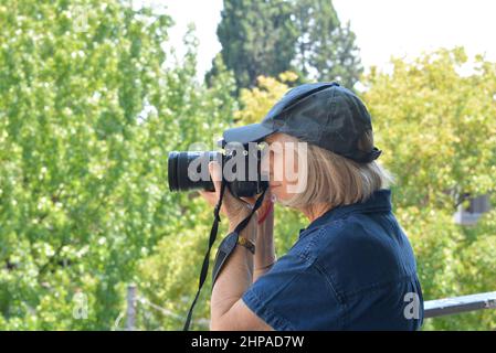 Photographe de femme âgée utilisant un appareil photo numérique reflex numérique au niveau des yeux. Robe bleue avec capuchon bleu sur un fond doux d'arbres Banque D'Images