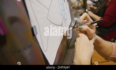 Une artiste apprend à peindre avec un aérographe avec du colorant acrylique, du papier et du chevalet. Intérieur. Concept art moderne, aérographe, aérographie, dessin pulvérisation image créer pulvérisateur dessin graffiti aérosol spray Banque D'Images
