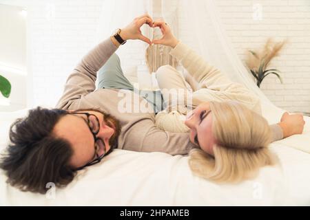 jeune couple caucasien se posant sur leur lit avec des feuilles blanches et mettant leurs mains ensemble créant un geste de coeur. Photo de haute qualité Banque D'Images