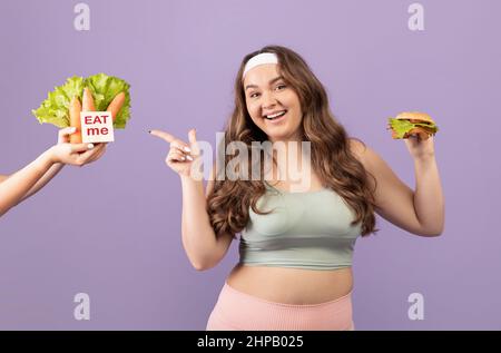 Bonne femme européenne millénaire de taille plus grande dans les vêtements de sport tenir le hamburger, en pointant le doigt sur les légumes offerts Banque D'Images