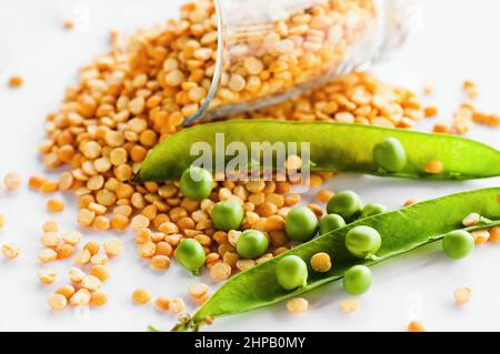 Petits pois verts frais en légumineuses ouvertes (et également gratuits) et petits pois jaunes séchés en verre et partiellement déversés d'un verre sur fond blanc Banque D'Images