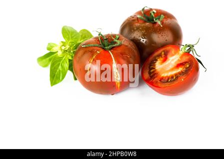 Trois tomates rouge foncé (variété kumato) avec feuille de basilic sur fond blanc, gros plan. Banque D'Images