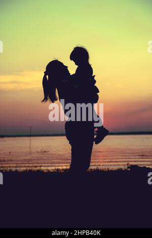 Silhouette de mère et d'enfant en profitant de la vue au bord de la rivière. Mère portant sa fille sur fond de ciel de coucher de soleil coloré. Famille sympathique. Croix Banque D'Images