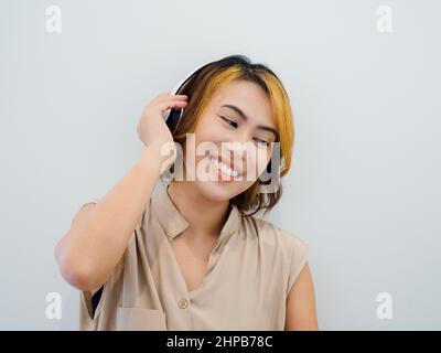 Portrait d'une femme asiatique heureuse avec cheveux courts en chemise sans manches beige Profitez de la musique, du podcast ou de la chanson à la radio avec un casque blanc sur W Banque D'Images