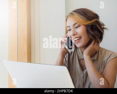 Le propriétaire d'affaires asiatique, la femme avec les cheveux courts tendance et l'air cool parler avec un partenaire d'affaires tout en travaillant avec l'ordinateur portable sur blanc W. Banque D'Images