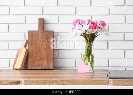 Vase avec fleurs et carte vierge sur le comptoir de cuisine près du mur en brique blanche Banque D'Images
