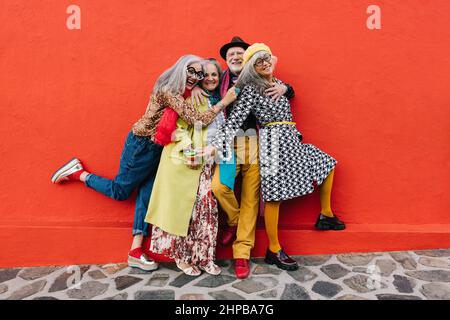 Groupe de personnes âgées excentriques qui rient et s'amusent tout en se tenant ensemble sur un fond rouge. Quatre amis âgés joyeux fêtant Banque D'Images