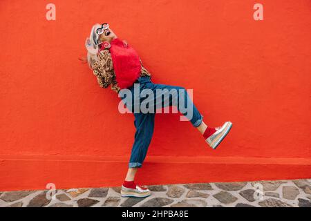 Femme âgée excitée dansant et s'amusant sur fond rouge. Heureuse femme âgée se sentant jeune et dynamique tout en portant des vêtements colorés Banque D'Images