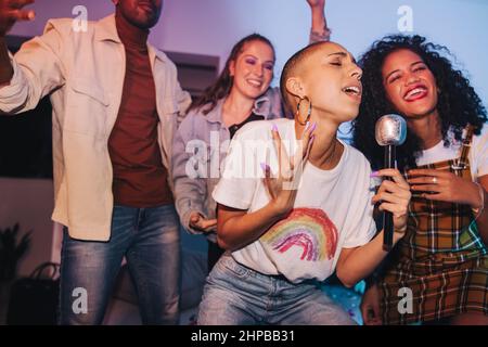Une jeune femme chantant son cœur pendant la soirée karaoké. Une jeune femme vibrante qui divertit ses amis avec un micro lors d'une fête à la maison. Groupe de Happy yo Banque D'Images