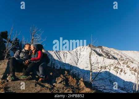 Homme et femme en vêtements de sport s'assoient sur la colline. Couple de voyageurs charmants à tenir, appréciant le paysage des hautes terres, beau coucher de soleil. Deux touristes font de la randonnée sur fond de montagnes enneigées. Banque D'Images