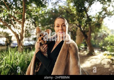 Femme de haut niveau regardant l'appareil photo tout en tenant son chiot à l'extérieur. Femme posh d'âge moyen debout seule dans un parc pendant la journée. Animal de compagnie femelle Banque D'Images