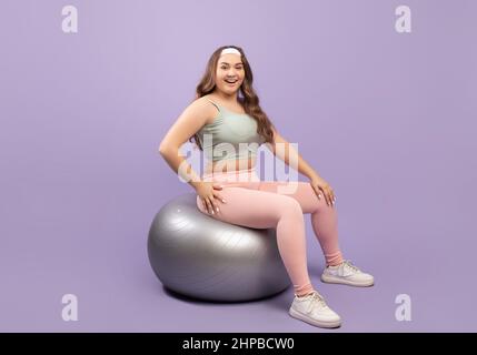 Joyeuse confiante et de taille plus européenne femme millénaire de sport assis sur le ballon de fitness isolé sur fond violet Banque D'Images