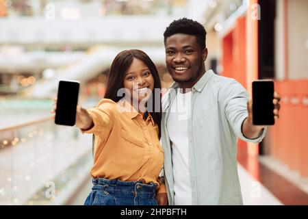 Heureux Noir couple montrant 2 smartphones avec écrans vides à l'appareil photo tenant deux gadgets dans les mains, les personnes présentant Free Copy Space pour maquette, Sele Banque D'Images