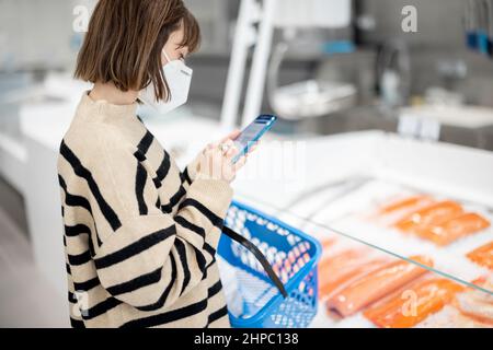 Femme dans le masque de visage en utilisant le téléphone tout en se tenant avec un panier à la boutique Banque D'Images