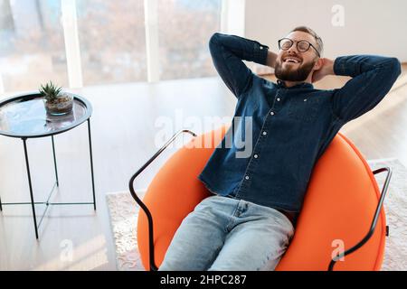 Joyeux gars à barbe avec les yeux fermés se reposer et se détendre assis dans la chaise en tenant les mains derrière la tête à la maison. Homme appréciant le week-end et le confort domestique Banque D'Images