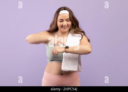 Joyeux heureux excitée plus grande taille femme européenne millénaire dans les vêtements de sport avec serviette de vérifier le pouls sur le tracker de forme physique isolé sur fond violet, studio. W Banque D'Images