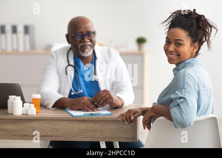 Portrait d'une femme afro-américaine souriante recevant une consultation médicale et regardant en arrière la caméra au premier plan, homme mature médecin sittin Banque D'Images