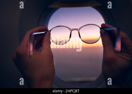 Main du passager tenant des lunettes contre la fenêtre de l'avion pendant le coucher du soleil coloré. Thèmes Voyage, vue et vision. Banque D'Images