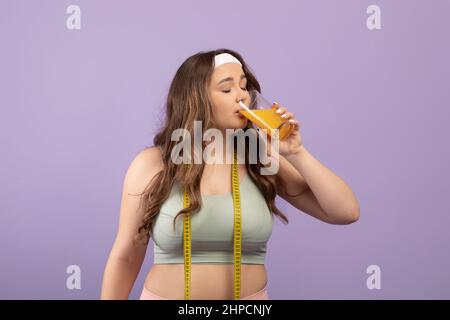 Calme jolie femme de taille plus jeune dans les vêtements de sport avec les yeux fermés et le ruban de mesure boire un verre de jus d'orange