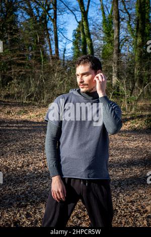Le coureur écoute de la musique tout en s'étirant dans les bois. Portrait d'un beau homme aux yeux bleus et d'une moustache qui s'entraîne dans le parc. Banque D'Images