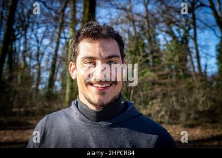 Le coureur écoute de la musique tout en s'étirant dans les bois. Portrait d'un beau sourire aux yeux bleus et d'une moustache qui s'entraîne dans le parc. Banque D'Images