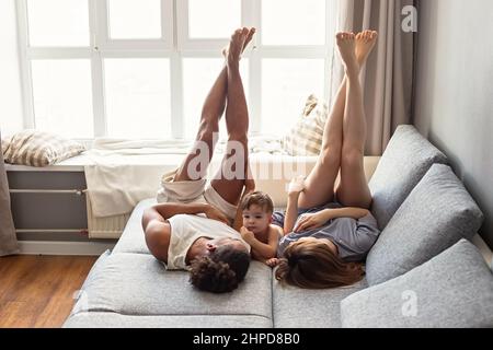 Une drôle de famille multiraciale avec bébé garçon , dans les vêtements confortables de la maison sont couchés sur le canapé près de la fenêtre Banque D'Images