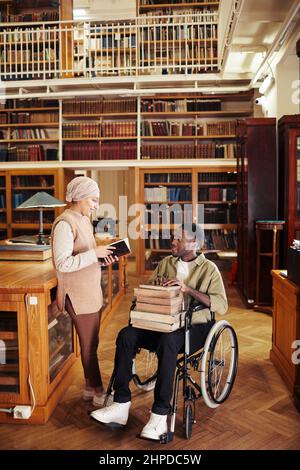 Portrait complet du jeune homme afro-américain en fauteuil roulant tenant une pile de livres et discutant avec un ami à la bibliothèque de l'université Banque D'Images