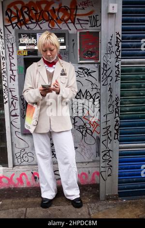 Une femme asiatique vérifie son téléphone tout en fume une cigarette. Derrière se trouve un cash point très gratitisé. Photo prise dans East Londons Brick Lane. Banque D'Images