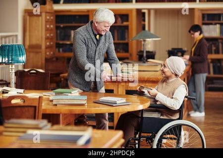 Portrait d'un professeur senior souriant aidant une jeune femme en fauteuil roulant à la bibliothèque de l'université Banque D'Images