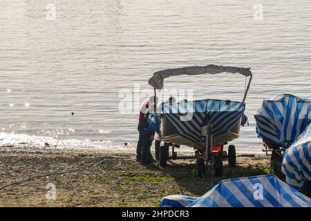 Vue en contre-jour d'une plage avec des personnes effectuant des travaux d'entretien sur un petit bateau dans une zone pour le stockage d'hiver, Sanremo, Imperia, Ligurie, Italie Banque D'Images