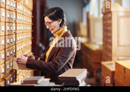 Vue latérale d'un bibliothécaire féminin recherchant un livre dans l'armoire du catalogue de la bibliothèque Banque D'Images