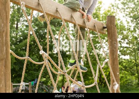 Un garçon actif jouant à l'extérieur, un filet de corde d'escalade pour enfant sur le terrain de jeu. Un enfant heureux qui profite d'une activité dans un parc d'escalade lors d'une journée d'été. Banque D'Images