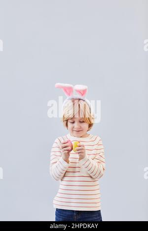 Portrait vertical à la taille d'un joli garçon blond portant des oreilles de lapin et jouant avec des œufs de Pâques sur fond blanc neutre, espace de copie Banque D'Images