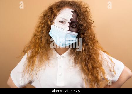 Portrait d'une belle fille à cheveux rouges avec des yeux bleus peints avec le drapeau du Qatar sur son visage a un masque médical sur son visage. Banque D'Images