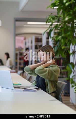 Femme fatiguée massant le cou et regardant la fenêtre tout en travaillant sur un ordinateur portable dans la bibliothèque publique Banque D'Images