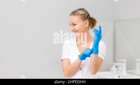 Une jeune femme souriante met des gants médicaux. Le cosmétologue prépare la procédure. Copier l'espace. Cosmétologie professionnelle. Banque D'Images