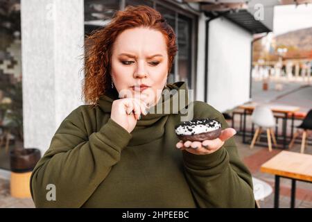 Portrait d'une femme jeune de taille plus grande tient un beignet de chocolat à la main. Le concept de régime alimentaire et de nutrition. Banque D'Images