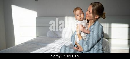 Une jeune femme s'assoit sur un lit et tient un bébé. Bonjour. Le concept de congé de maternité et de maternité. Defocus Banque D'Images