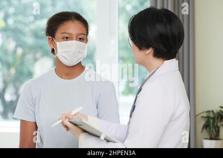 Médecin asiatique en manteau blanc parlant à une fille dans le masque et remplissant la carte médicale pendant leur conversation à l'hôpital Banque D'Images