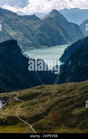 Vue aérienne d'un paysage alpin avec un sentier qui s'enroule à travers les pâturages. Sur le sentier sont deux personnes marchant vers quelques vaches. En arrière-plan se trouve un lac de montagne, vers lequel le sentier se dirige. Banque D'Images