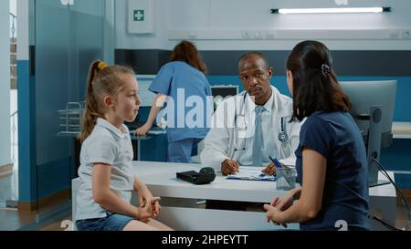 Spécialiste de la santé discutant avec l'enfant du rétablissement de la maladie et prenant des notes sur l'ordinateur pour donner le traitement. Médecin ayant une conversation avec une petite fille et une mère lors d'une visite de consultation. Banque D'Images