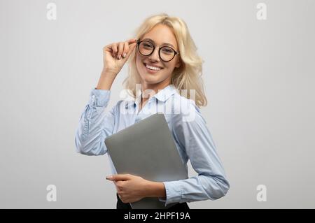 Jeune femme d'affaires réussie en tenue formelle tenant un ordinateur portable et touchant des lunettes, souriant à l'appareil photo Banque D'Images