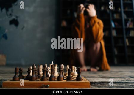 Gros plan sélectif sur le jeu d'échecs sur parquet, inquiet femme méconnaissable assise loin près des étagères de la bibliothèque sombre. Banque D'Images