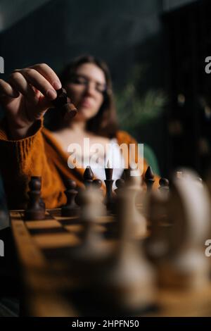 Mise au point verticale sélective d'une femme d'affaires confiante dans des lunettes élégantes faisant bouger les échecs assis sur un fauteuil dans une pièce sombre. Banque D'Images