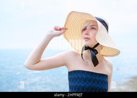 Caucasien belle blonde élégante triste femme en robe bleue et en chapeau de paille debout près de la mer Banque D'Images