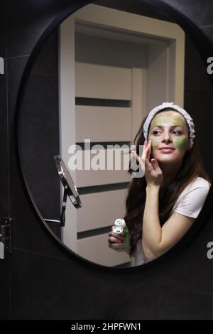 Fille prenant soin de teint huileux. Femme en bandeau appliquant un masque de boue d'argile verte sur son visage dans une salle de bains intérieure sombre à la maison devant un miroir. Banque D'Images