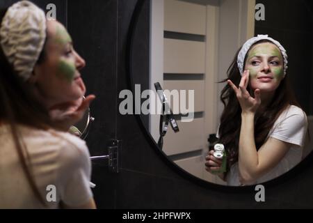 Femme en bandeau appliquant un masque en argile verte sur son visage dans une salle de bains intérieure sombre devant un miroir. Fille prenant soin de teint huileux. Natura Banque D'Images